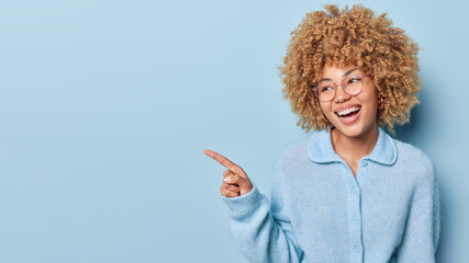 Studio shot of pretty curly haired woman points index finger on empty space for your advertising...
