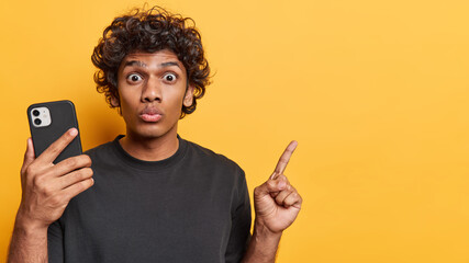 Horizontal shot of curly haired Hindu man with stunned expression holds modern smartphone points...