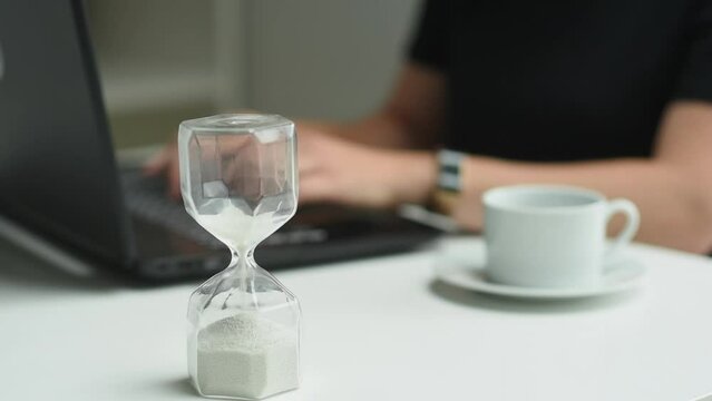 woman is typing on the keyboard, the hourglass time is running out and the woman closes the laptop. Time is up. An hourglass is depicted in the foreground, the background is blurred. Study, business, 