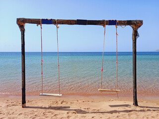 Rustic swing on the beach sand, sea and turquoise water under blue sky of the Dahab desert. Wooden and rope swing. Games and outdoor fun.