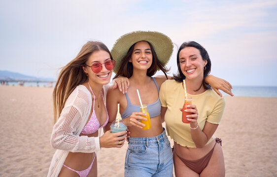 Happy portrait of three young Caucasian women on beach together hugging looking at camera cheerful. Smiling girls friends holding fruit smoothies enjoying their summer vacation in Mediterranean.