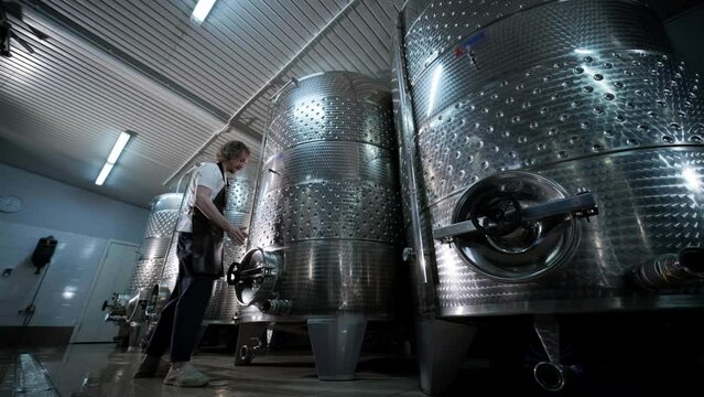 A happy successful farmer or winemaker is walking in the middle of wine tanks and checking a fermentation grade of high quality wine production in a modern winemaking facility or winery.
