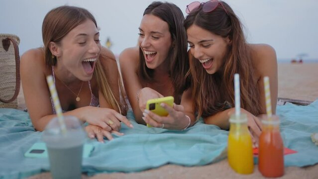 Group of three smiling young women using mobile phone app lying on beach. Cheerful girls enjoying weekend free time. Concept of summer vacation, friendship and social networking in generation z.