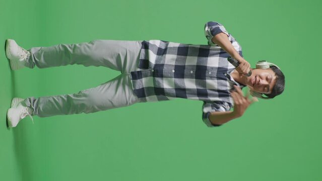 Full Body Of Young Asian Teen Boy Wearing Headphone And Holding A Microphone And Rapping On The The Green Screen Background
