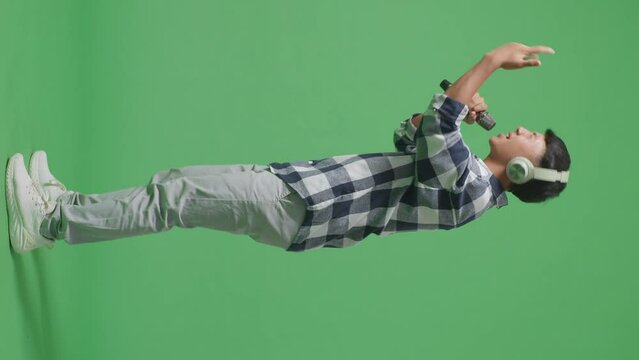 Full Body Side View Of Young Asian Teen Boy Wearing Headphone And Holding A Microphone And Rapping On The The Green Screen Background

