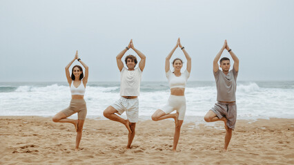 Healthy lifestyle concept. Group of men and women practising yoga, standing in tree pose on beach