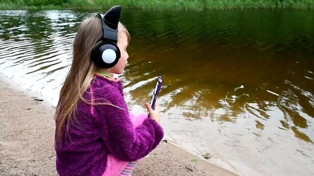A little charming girl with long hair sits on the shore of the lake. Listens to music in headphones in the shape of cat ears.