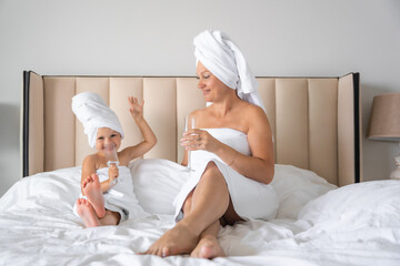 Happy family mother and little daughter in dressing gowns and towels laugh and drinking water with glasses during spa day at home