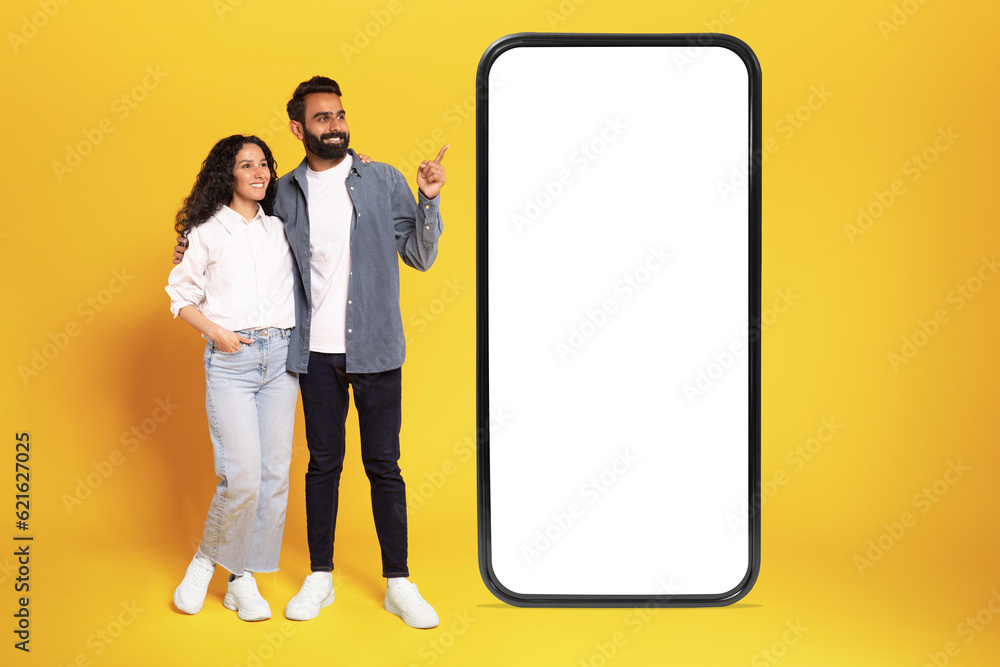 Wall mural Happy arab couple showing smartphone screen recommending application, yellow background