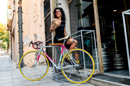Young ethnic woman sitting on stool with bicycle on street and drinking hot coffee