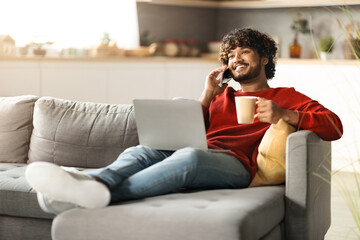 Indian Guy Talking On Cellphone, Relaxing On Couch With Laptop And Coffee