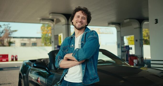 A young brunette guy in a denim jacket poses and looks at the camera, he folded his arms on his chest and leaned on his dark gray Convertible at a gas station