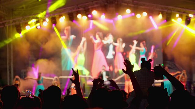 Happy people watching amazing musical concert. Bright colorful stage lighting. Nightlife and entertainment concept. People with raised arms on music event with lights. 4K, UHD