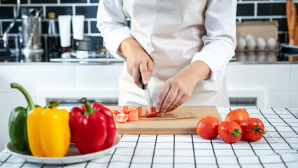 Asian housewife wearing apron and using knife to slice tomato on chopping board while preparing ingredient