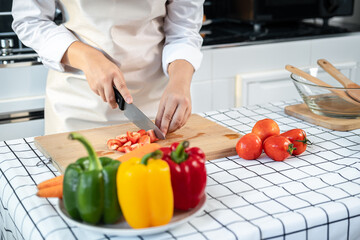 Asian housewife wearing apron and using knife to slice tomato on chopping board while preparing ingredient