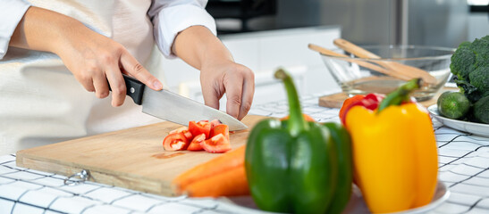 Close up of asian housewife wearing apron and using knife to slice tomato on chopping board while...