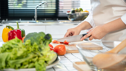 Obraz na płótnie Canvas Asian housewife wearing apron and using knife to slice tomato on chopping board while preparing ingredient