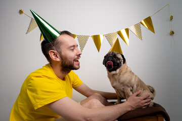 Caucasian man owner congratulation cute funny pug dog with pet birthday in a party hat. Domestic animal love and pampering concept. Celebrating pets birthday enjoys