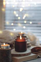 Cozy autumn or winter composition with aromatic candle, wool sweater, fairy lights, book. Aromatherapy, home atmosphere of cosiness and relax. Wooden background close up