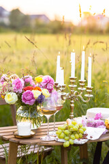 Dinner table for outdoor wedding or romantic date for a couple in a field. Elegant decoration with vintage chandelier, ranunculi flowers in vase, light snacks, cheese, fruits, white wine.