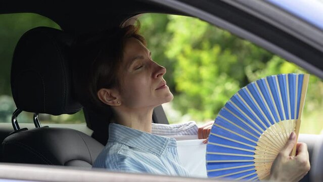 Tired exhausted middle aged woman waving blue fan suffers from stuffiness driving car on summer hot weather. Overheating, sultriness, high temperature, swelter in car with broken air conditioner.