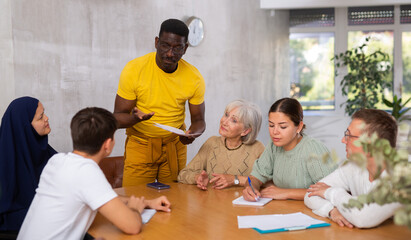 Focused african american man conducting informal meeting with international mixed age group of colleagues sitting around table in office, discussing new strategies, projects, or work-related problems