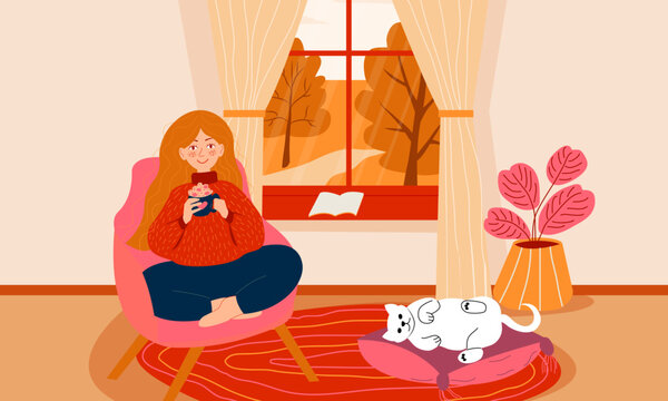 Woman sitting in a chair in front of window at home with coffee in hands, cat is laying. Autumn landscape outside the window