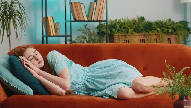 Tired young redhead woman lying down in bed taking a rest at home. Portrait of carefree girl napping, falling asleep on comfortable orange sofa with pillows. Closed her eyes enjoy daytime nap alone