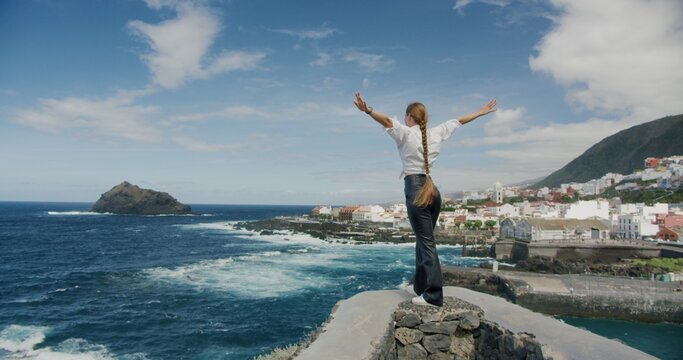 Lonely girl spreads her arms looks at the Atlantic Ocean. Woman with long blond braid hair stands with her back to camera. Tenerife. Gorachico.