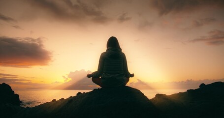 Silhouette of relaxed woman against sunset sky on rocky beach at sunset. Girl meditates in nature and sun rays illuminate her.