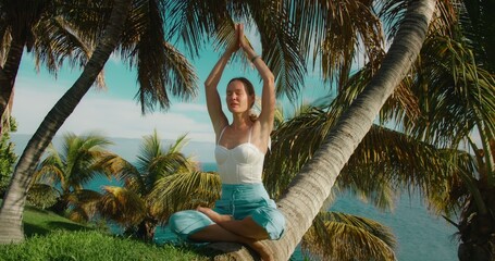 Girl sits on a lush grassy lawn in a lotus position and breathes deeply calmly. Beautiful tropical...