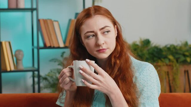 Attractive smiling young redhead woman drinking a cup of warm coffee or herbal tea sitting at home living room couch in the morning. Lovely girl enjoying comfortable relaxing, having a break alone