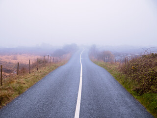 Asphalt road and fields in country side in a fog. Dangerous driving conditions with low visibility and wet road surface. Mist over wild nature. Irish country side.