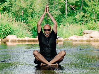 Bald male in dark wet shirt and shorts sitting in a stream and practice yoga relaxation. Selective...