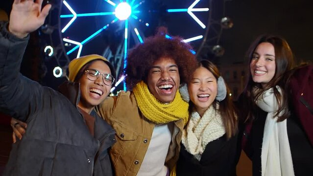 Group of multiethnic friends having fun in a city street during a winter night illuminated at Christmas. Friends against illuminated ferris wheel outdoor.