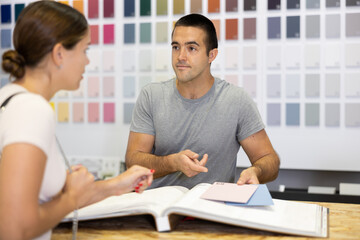 Obraz premium Male salesperson in paint and varnish department is assisting female customer by showcasing product samples and providing guidance through product catalog at counter.