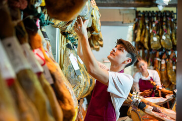 Young man in apron working in jamoneria, arranging pig's legs at counter.