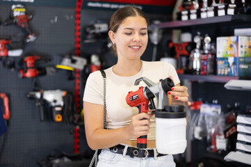 Smiling young woman involved in crafts or hobbies using of paint, choosing air sprayer in local hardware store - obrazy, fototapety, plakaty