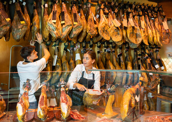 Cheerful caucasian young man and woman in uniform carving jamon slices from dry-cured pig's leg. Jamoneria workers cutting ham with knife.
