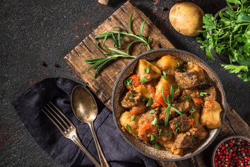 Beef meat stewed with potatoes, herbs and carrots in a plate with a spoon on black background. top...