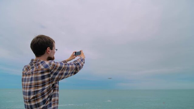 Man holds smartphone, takes photo or shoots video of arriving passenger airplane, airliner against the overcast sky - back view, wide angle view. Photography, plane spotting and technology concept