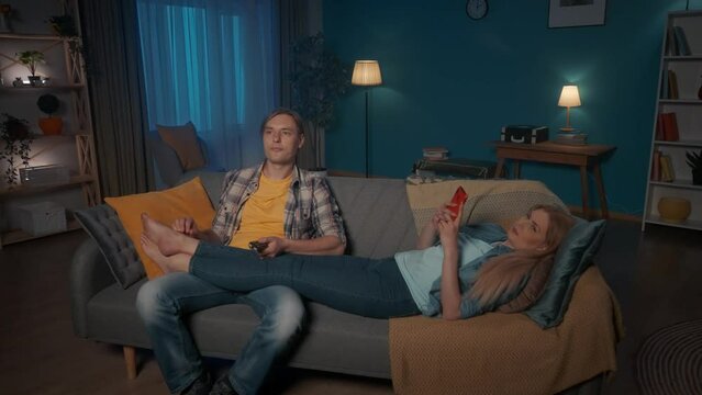 A married couple is relaxing in the evening. A man sits on the couch watching TV, switching channels using the remote control. The woman lies with the phone, putting her legs on the man's knees.
