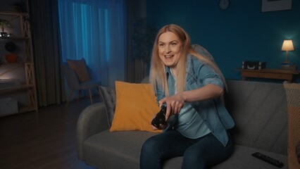 A woman plays a consular game with a game controller while sitting on the couch at home in the evening. Concentrated woman presses joystick, nervous.