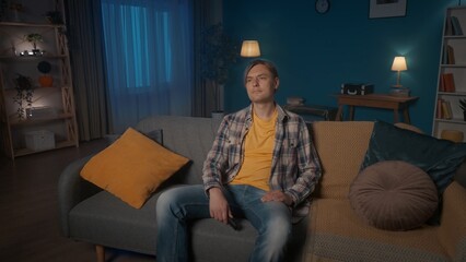 Fototapeta premium A young man sits on the sofa in the evening in the room and watches TV. A man is sitting with a TV remote control.