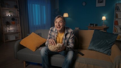 A man sits on the couch in the evening, eats popcorn and watches TV. A man watches a comedy, a comedy show, laughs happily.