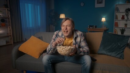 Obraz premium A man sits on the couch in the evening, eats popcorn and watches TV. A man watches a comedy, a comedy show, laughs happily.