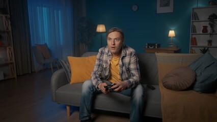 Obraz premium A man plays a consular game with a game controller while sitting on the couch at home in the evening. A man often presses the joystick, gets nervous.