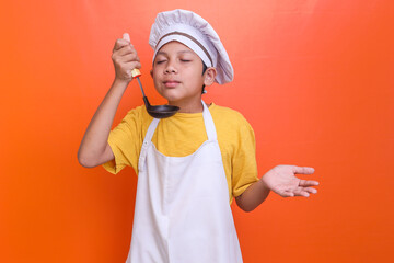 Portrait of little boy cook holding a soup ladle and smelling it with his eyes closed while...