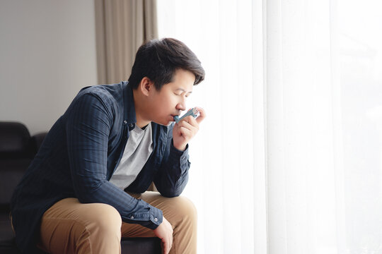 Sick Asian young man using inhaler to treat asthma and respiratory diseases at home. Asthma attack. Concept of allergy care.