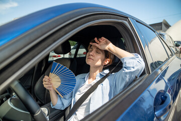 Exhausted tired middle aged woman drives car waves blue fan suffers from stuffiness stands in urban...